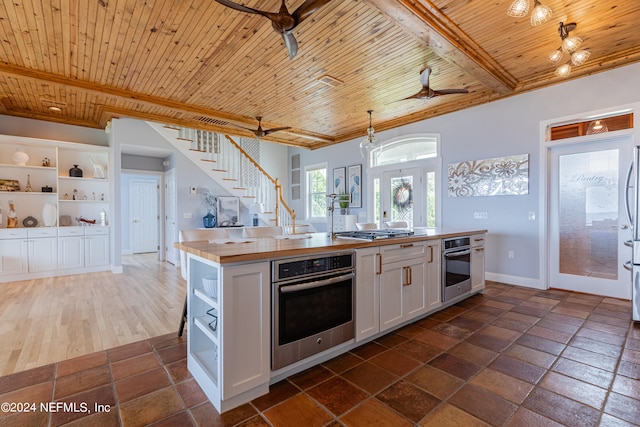 kitchen with appliances with stainless steel finishes, ceiling fan, beam ceiling, a kitchen island with sink, and dark tile floors