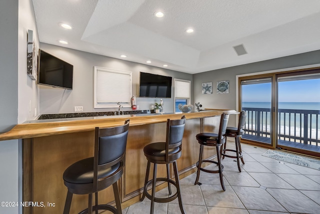 bar featuring light tile floors, a textured ceiling, a water view, a tray ceiling, and butcher block counters