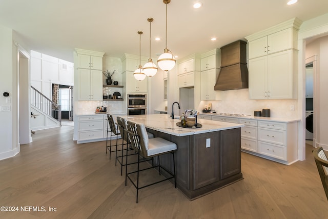 kitchen with pendant lighting, white cabinetry, light stone counters, custom range hood, and a center island with sink