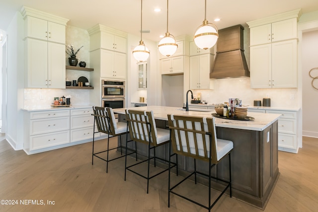 kitchen featuring double oven, decorative light fixtures, a breakfast bar area, a kitchen island with sink, and custom range hood