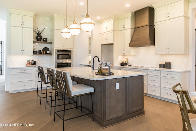 kitchen featuring pendant lighting, double oven, light stone countertops, custom range hood, and a center island with sink