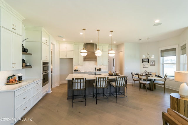 kitchen featuring premium range hood, decorative light fixtures, hardwood / wood-style flooring, a kitchen island with sink, and white cabinets