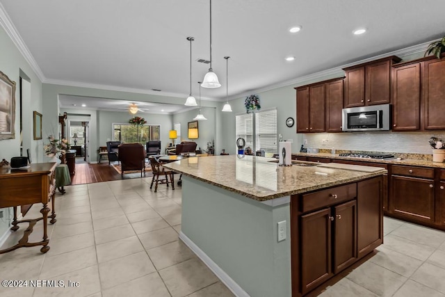 kitchen with an island with sink, ceiling fan, light stone counters, hanging light fixtures, and appliances with stainless steel finishes