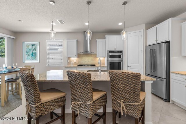 kitchen featuring light stone countertops, a kitchen island with sink, appliances with stainless steel finishes, light tile floors, and wall chimney exhaust hood