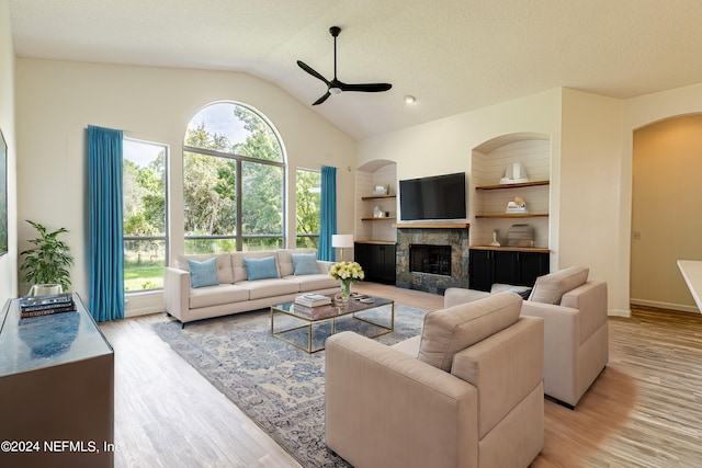 living room featuring lofted ceiling, a stone fireplace, light hardwood / wood-style flooring, a textured ceiling, and ceiling fan