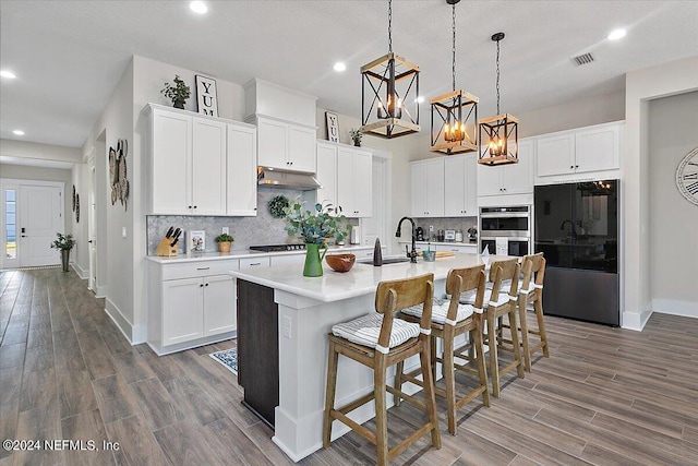 kitchen with black refrigerator, decorative light fixtures, stainless steel double oven, backsplash, and a center island with sink