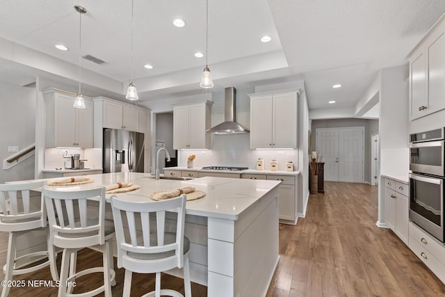 kitchen with wood finished floors, a tray ceiling, a sink, appliances with stainless steel finishes, and wall chimney range hood