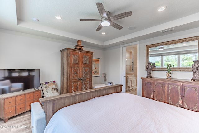 bedroom with a raised ceiling, ceiling fan, ensuite bathroom, and light hardwood / wood-style flooring