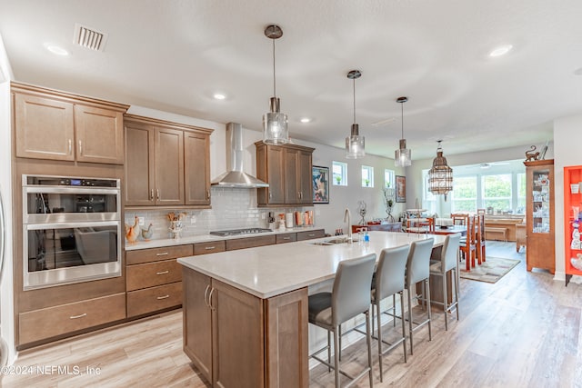 kitchen with a kitchen island with sink, a breakfast bar, light hardwood / wood-style floors, stainless steel appliances, and wall chimney exhaust hood