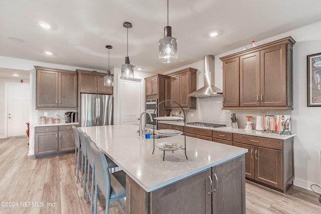 kitchen featuring decorative light fixtures, appliances with stainless steel finishes, a kitchen island with sink, backsplash, and wall chimney range hood