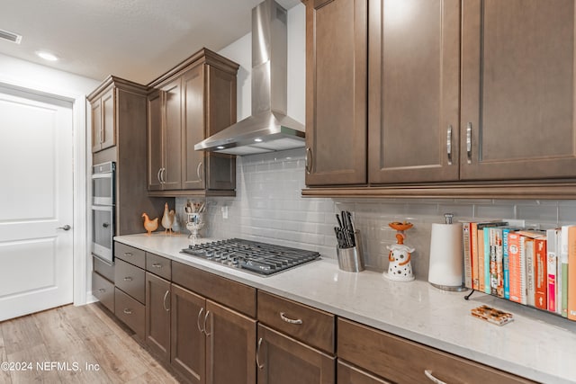 kitchen with appliances with stainless steel finishes, light wood-type flooring, backsplash, light stone countertops, and wall chimney exhaust hood