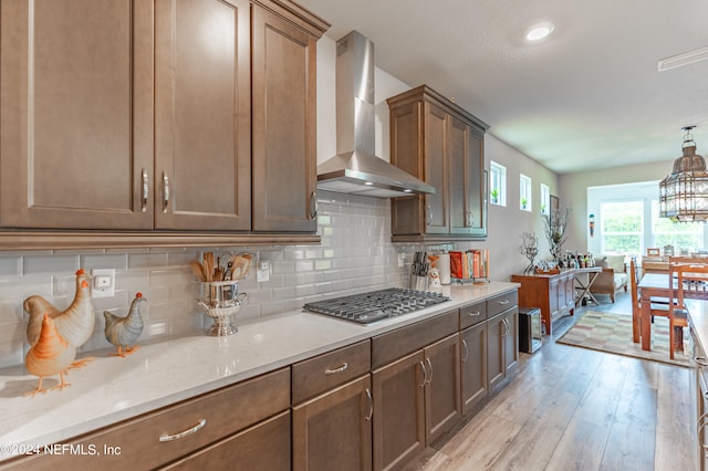 kitchen featuring pendant lighting, wall chimney range hood, stainless steel gas stovetop, tasteful backsplash, and light hardwood / wood-style flooring