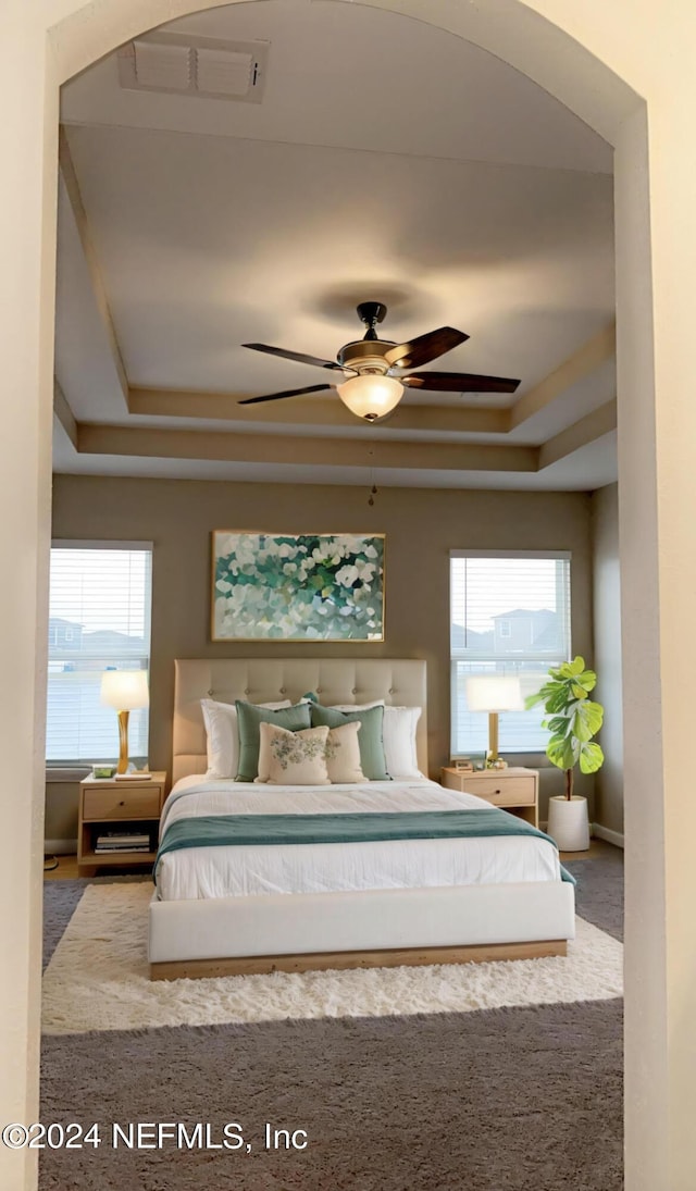 carpeted bedroom with ceiling fan, a tray ceiling, multiple windows, and visible vents