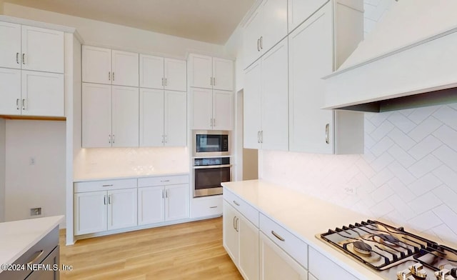 kitchen featuring appliances with stainless steel finishes, decorative backsplash, light hardwood / wood-style floors, white cabinetry, and custom exhaust hood