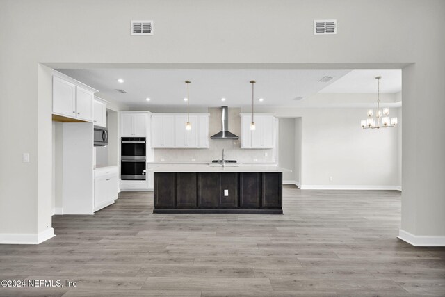 kitchen with wall chimney range hood, light wood-type flooring, backsplash, a kitchen island with sink, and appliances with stainless steel finishes