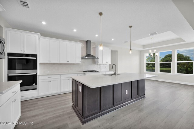 kitchen with light hardwood / wood-style flooring, tasteful backsplash, stainless steel double oven, a raised ceiling, and wall chimney range hood