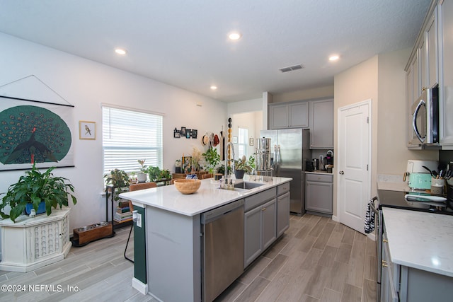 kitchen with an island with sink, sink, gray cabinets, stainless steel appliances, and light wood-type flooring