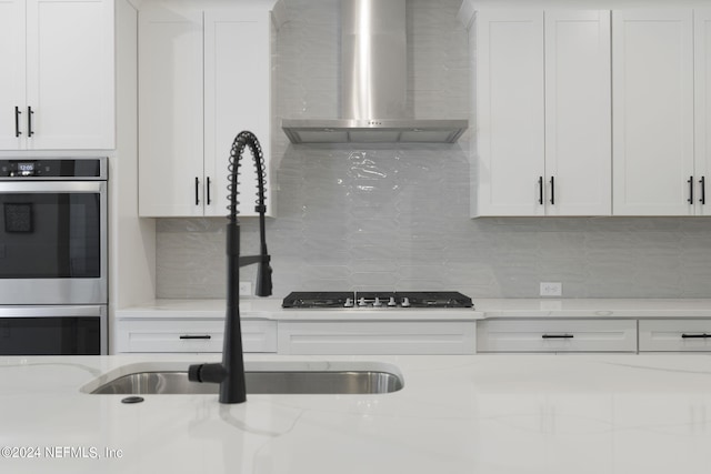 kitchen with white cabinets, light stone countertops, stainless steel double oven, wall chimney range hood, and backsplash