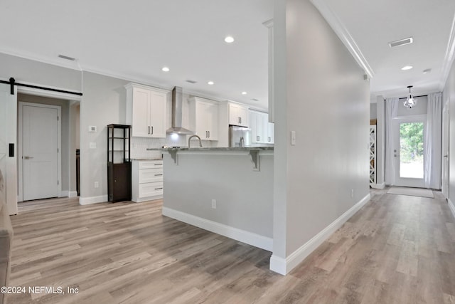 kitchen with wall chimney exhaust hood, a barn door, crown molding, a breakfast bar, and white cabinets