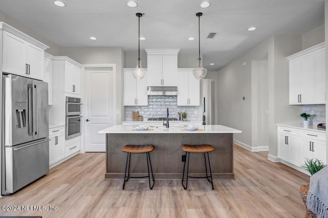 kitchen with light wood-type flooring, stainless steel appliances, a kitchen island with sink, white cabinets, and hanging light fixtures