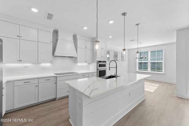 kitchen featuring sink, hanging light fixtures, light hardwood / wood-style floors, a center island with sink, and custom exhaust hood