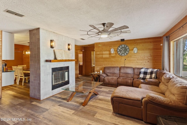 living room with wood walls, a large fireplace, light hardwood / wood-style flooring, ceiling fan, and a textured ceiling