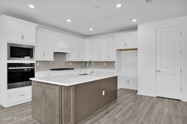 kitchen with black oven, light hardwood / wood-style flooring, sink, an island with sink, and stainless steel microwave