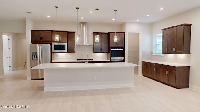 kitchen with appliances with stainless steel finishes, dark brown cabinets, wall chimney range hood, pendant lighting, and an island with sink