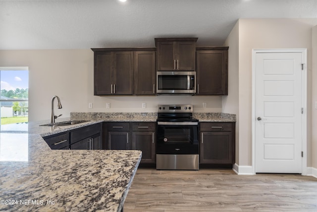 kitchen featuring light stone counters, stainless steel appliances, a sink, dark brown cabinets, and light wood-type flooring