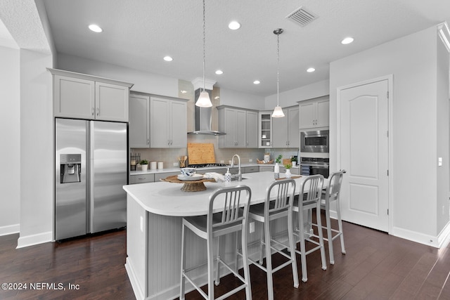 kitchen with a kitchen island with sink, built in appliances, wall chimney exhaust hood, gray cabinets, and decorative light fixtures