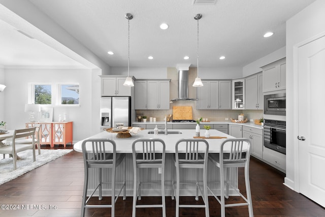 kitchen featuring pendant lighting, gray cabinetry, a center island with sink, wall chimney exhaust hood, and appliances with stainless steel finishes