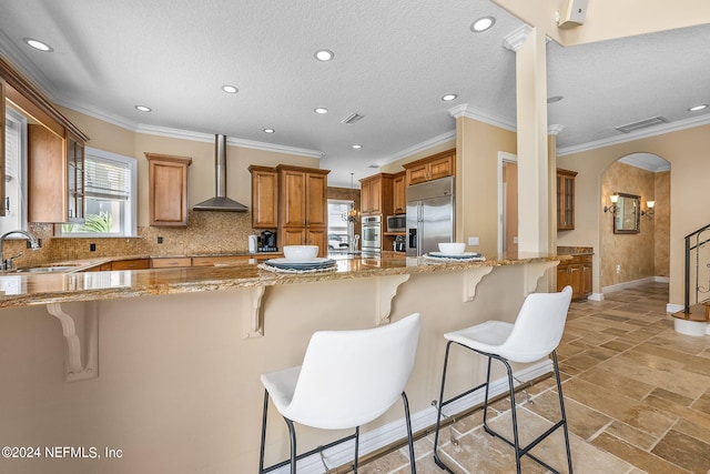 kitchen featuring sink, wall chimney range hood, light stone counters, built in appliances, and a breakfast bar