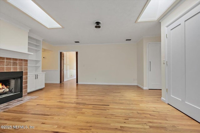 unfurnished living room featuring a tiled fireplace, a skylight, built in features, and light hardwood / wood-style flooring