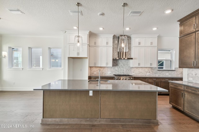 kitchen with sink, wall chimney range hood, hardwood / wood-style floors, an island with sink, and decorative light fixtures