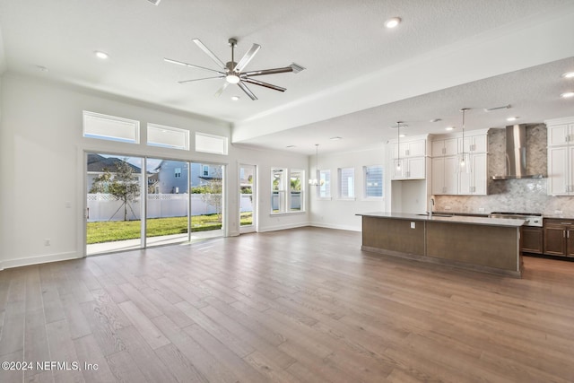 kitchen with sink, white cabinetry, wall chimney range hood, a kitchen island with sink, and backsplash