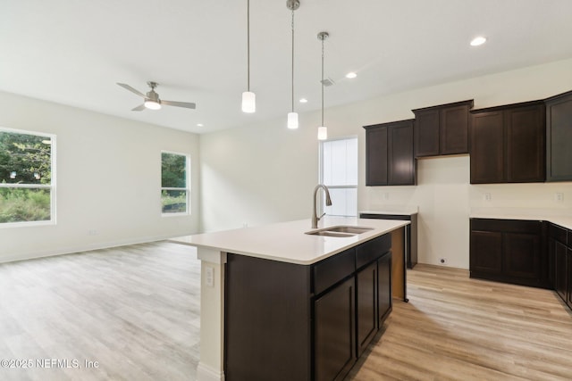 kitchen featuring ceiling fan, sink, hanging light fixtures, an island with sink, and light wood-type flooring