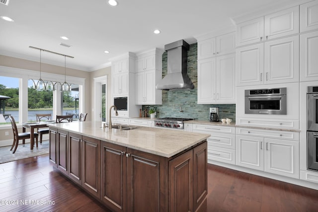 kitchen with wall chimney exhaust hood, sink, white cabinetry, decorative light fixtures, and a kitchen island with sink