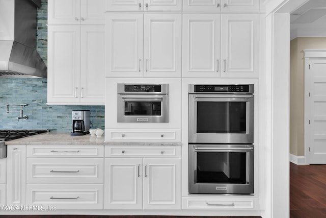 kitchen with double oven, light stone counters, white cabinets, decorative backsplash, and wall chimney exhaust hood