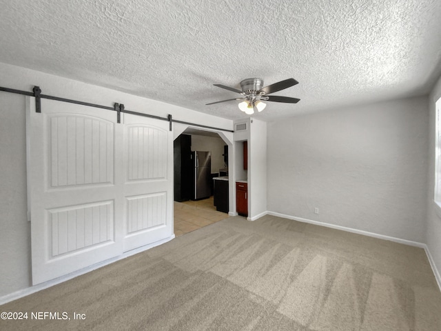 unfurnished bedroom featuring ceiling fan, stainless steel fridge, a barn door, a textured ceiling, and light colored carpet