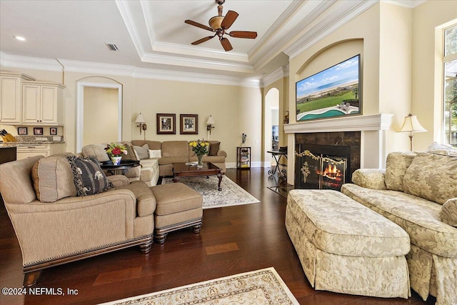 living room with ceiling fan, a tray ceiling, dark wood-type flooring, a tile fireplace, and ornamental molding