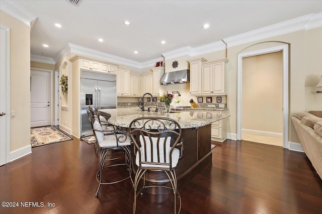 kitchen featuring a center island with sink, dark wood-type flooring, light stone counters, wall chimney exhaust hood, and built in refrigerator