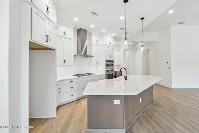 kitchen with wall chimney exhaust hood, a kitchen island with sink, pendant lighting, light hardwood / wood-style flooring, and white cabinetry