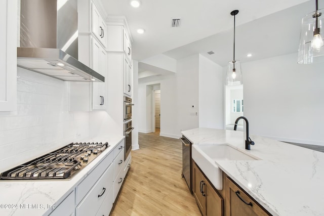 kitchen featuring white cabinets, wall chimney exhaust hood, light wood-type flooring, appliances with stainless steel finishes, and decorative light fixtures