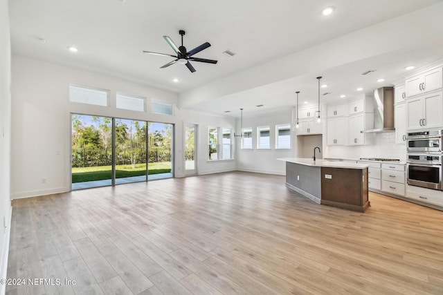 kitchen with wall chimney range hood, light hardwood / wood-style flooring, pendant lighting, a center island with sink, and white cabinets