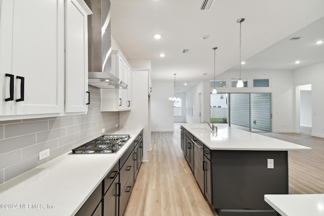 kitchen featuring hanging light fixtures, wall chimney range hood, stainless steel gas cooktop, light hardwood / wood-style flooring, and a kitchen island with sink