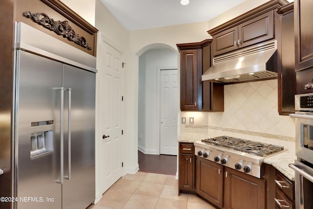 kitchen with ventilation hood, stainless steel appliances, light stone countertops, arched walkways, and backsplash