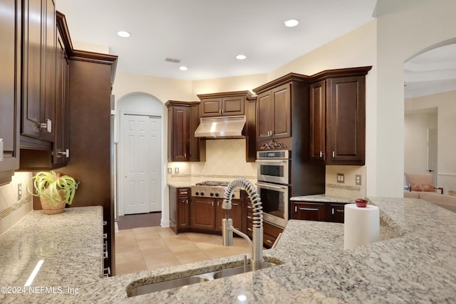 kitchen with light stone counters, a sink, under cabinet range hood, dark brown cabinetry, and double oven