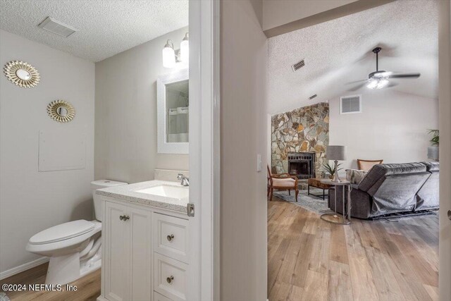 bathroom with a textured ceiling, wood-type flooring, a stone fireplace, large vanity, and toilet