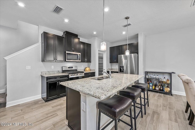 kitchen featuring hanging light fixtures, light hardwood / wood-style flooring, appliances with stainless steel finishes, light stone countertops, and a kitchen island with sink