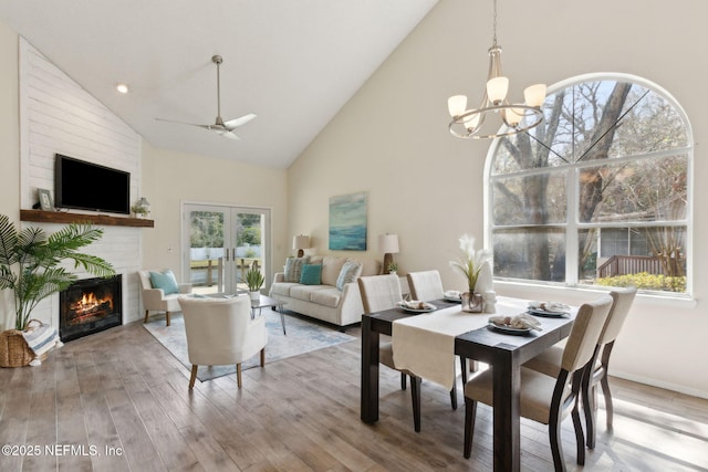 dining area featuring wood finished floors, high vaulted ceiling, french doors, a glass covered fireplace, and ceiling fan with notable chandelier